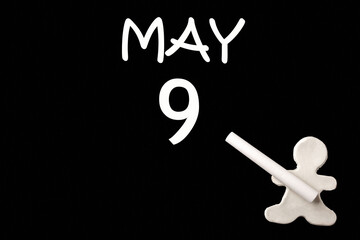 A small white plasticine man writing the date 9 May on a black board. Business concept. Education concept.