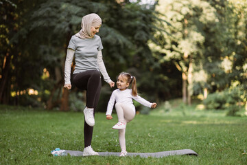 Yoga and stretching exercises of mom with child outdoors. Likable young Muslim mother and her cute daughter, having outdoor workout in the park, standing on yoga mat and doing high stepping exercise - Powered by Adobe
