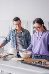 Teen girl with down syndrome holding flour near ingredients and friend in kitchen.