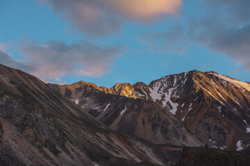 Scenic landscape with high snow mountain with sharp rocky pinnacle in golden sunlight under clouds of sunset color at changeable weather. Colorful view to large mountain top under orange clouds in sky