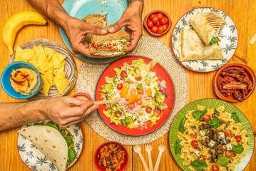 Italian and Mexican fast food dishes and appetizers, pasta salad, stuffed burrito, hummus with tortilla chips, vegetable sandwich, hands holding food and fruit salad