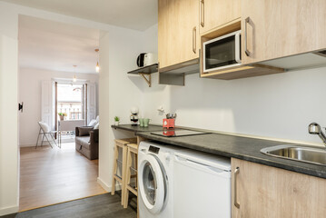 Kitchen with dark gray wooden countertops, wooden furniture and small appliances on the counter and access to a living room with sofas and white folding tables
