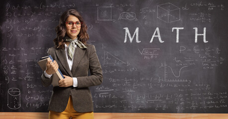 Young female math male teacher holding books and posing in front of a blackboard with formulas