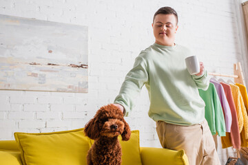 Teenage boy with down syndrome holding cup and petting poodle on couch.