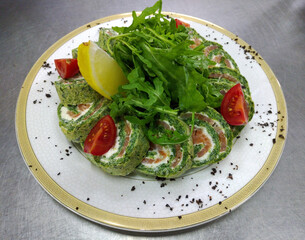 spinach roll with cream cheese and salmon on a round white plate. garnished with cherry tomatoes, arugula and lemon wedges
