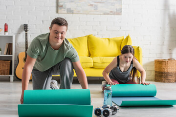 Smiling teen boy with down syndrome holding fitness mat near dumbbells and friend at home.