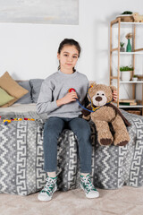 girl looking at camera while playing with teddy bear and toy stethoscope in bedroom.