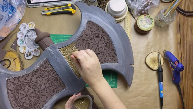 Woman makes viking axe for cosplay festival. Girl draw ornamental hatchet with gold paint and brush. DIY celtic weapons in craft workshop.
