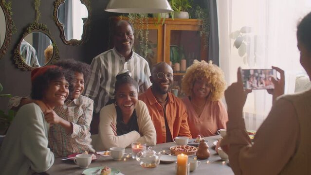 Large African American family smiling and posing together on camera at dinner table as girl taking picture with smartphone during home celebration