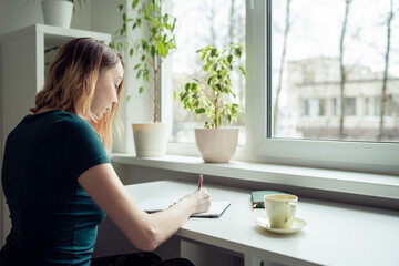 Young woman sitting at the table by the window and writing.