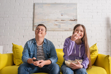 Positive teenager with down syndrome holding popcorn near friend with remote controller at home.