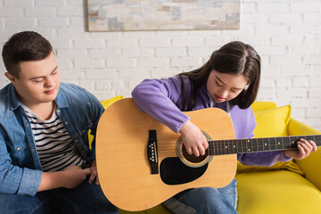 Girl with down syndrome playing acoustic guitar near friend on couch at home.