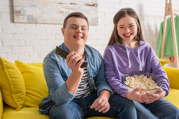 Happy teenagers with down syndrome holding remote controller and popcorn on couch.