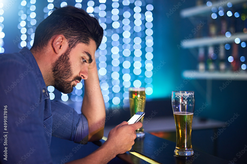 Poster drunken texts are never a good idea. shot of a young man looking upset while using his phone at a ba