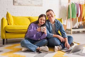 Positive couple of teenagers with down syndrome holding cup and using laptop at home.