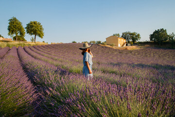 The beautiful young girl in a blue dress and cap walks across the field of a lavender, long curly hair, smile, pleasure, a house of the gardener in the background, trees, perspective of a lavender