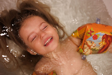 Little 3-year-old baby girl taking bath in tub at home. Portrait of happy child with long blond...