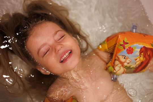 Little 3-year-old baby girl taking bath in tub at home. Portrait of happy child with long blond hair swimming in bath. Funny beautiful little girl smiling, smimming with armbands