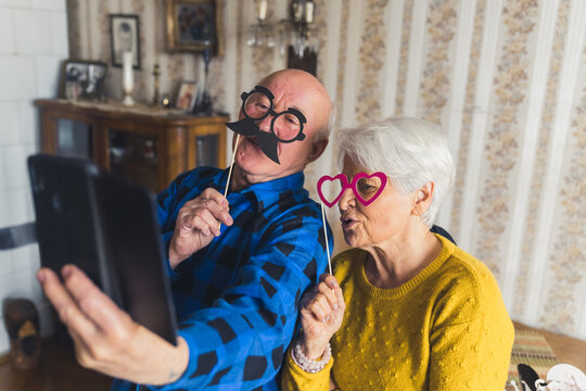 Playful and cheerful elderly couple takes a selfie in their vintage living room with props like fake heart-shaped glasses and mustache. High quality photo