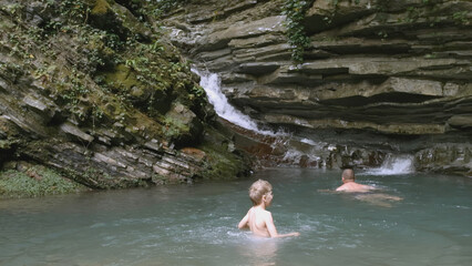 The boy enters the water near the waterfall to swim. CREATIVE. A white child in blue shorts enters the water. In the mountains there is a guy waist-deep in water