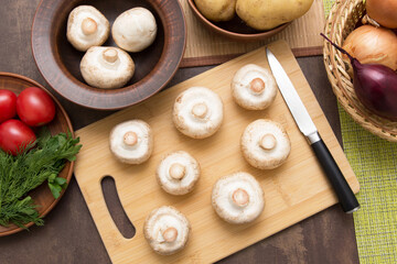 Mushrooms champignon on wooden cutting board with different vegetables on kitchen. Cooking vegetarian vegan diet dish, food. Top view, flat lay	

