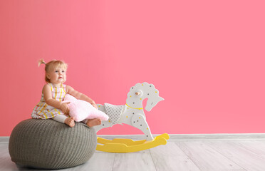 Adorable baby girl with pillow sitting on pouf near rocking horse