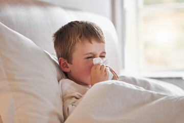 Achooo. Shot of a little boy feeling ill in bed at home and blowing his nose.