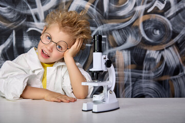Young laboratory worker rested his head on his hand, sitting in biology class table with microscope. Cute boy with glasses dreams of becoming great scientist and will make many different discoveries