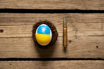 One white chicken egg with a painted yellow-blue flag of Ukraine in a straw basket and one bullet,...