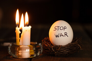 Stop war is written on a chicken white egg that lies next to a burning candle in the dark in...