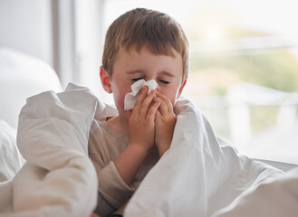 My nose wont stop dripping. Shot of a little boy feeling ill in bed at home and blowing his nose. - obrazy, fototapety, plakaty