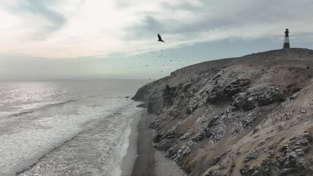 Aerial shot of the waves breaking on the cliffs with sea birds and a lighthouse.Half Moon beach, Puerto Eten, Lambayeque, Peru.