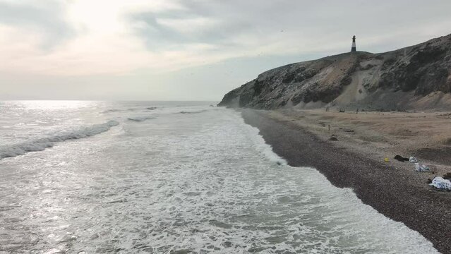 Aerial view of the breaking waves on the rocky coast with a lighthouse, birds and cliffs in the background. Media Luna beach, Puerto Eten, Lambayeque, Peru.