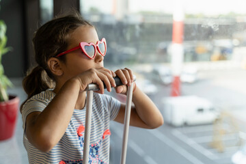 little girl with suitcase and glasses at the airport is waiting for boarding near a large window....