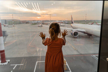 Little girl at the airport is waiting for landing at the big window. Cute kid stands at the window...