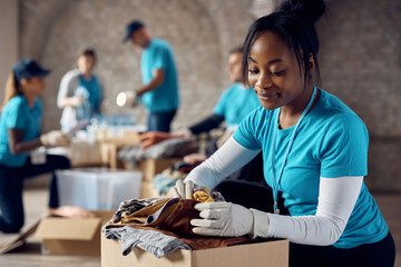 Black female volunteer packing clothes in donation boxes while working at community center. - obrazy, fototapety, plakaty