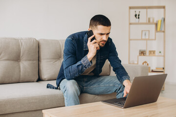 Portrait of smiling guy talking on mobile phone, working on laptop, sitting on the sofa in living room.