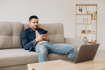 Portrait of smiling guy talking on mobile phone, working on laptop, sitting on the sofa in living room.