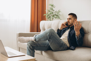 Fototapeta premium Portrait of smiling guy talking on mobile phone, working on laptop, sitting on the sofa in living room.