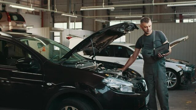 Handsome mechanic in uniform is using a laptop while repairing car in auto service. Auto mechanic fixing a vehicle in service station. Car mechanic working at automotive service center.