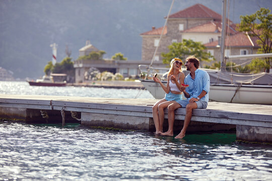 Happy couple laughing together and enjoying a piece of watermelon, sitting by water on wooden jetty. Couple in love. Tourism, summertime, togetherness, lifestyle concept.