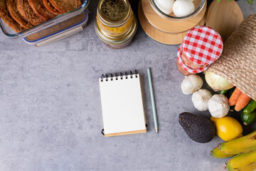 Fototapeta na wymiar Blank spiral notepad with a pencil beside it surrounded by a lot of prepared food inside glass jars over a kitchen table. Horizontal photo.