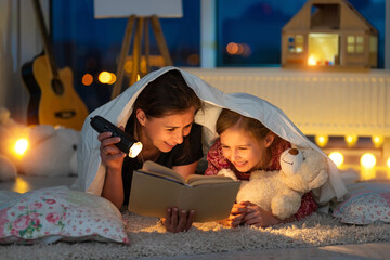 Happy mother with little daughter reading book using flashlight under blanket on floor in...