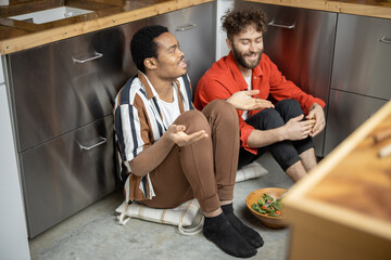 Two brightly dressed stylish guys having close conversation, sitting together on a kitchen floor and eating healthy salad. Concept of gay relations. Caucasian and latin man bonding while talking close