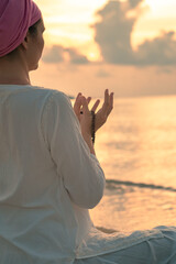 Close up woman in white clothes and a turban sits by the sea on a sandy beach at dawn on a summer morning and doing yoga exercises . Colorful sky on the background.