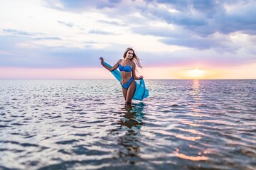 A girl in a blue swimsuit and a bright pareo posing against the background of a sunset in estuary with transparent water