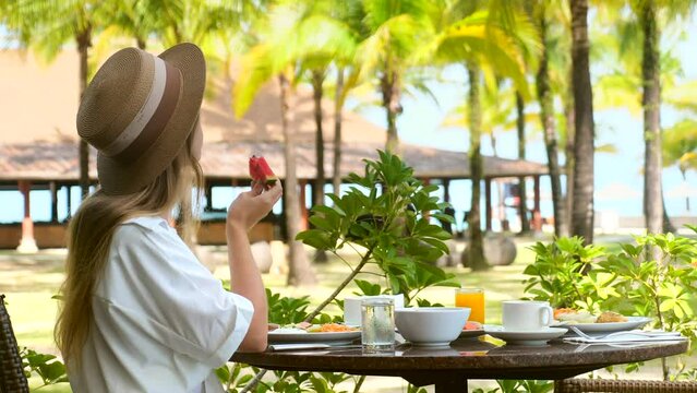 Young woman eat fresh fruits in outdoor hotel restaurant. Girl have healthy breakfast on terrace of cafe of luxury resort on tropical island among greenery, palm trees with sea view. Vacations concept
