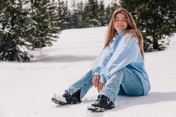 A young, beautiful girl in the mountains sits in the snow among the trees