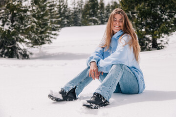 A young, beautiful girl in the mountains sits in the snow among the trees