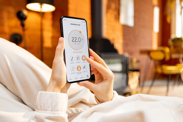 Woman holding phone with running smart home application for heating temperature control, while...